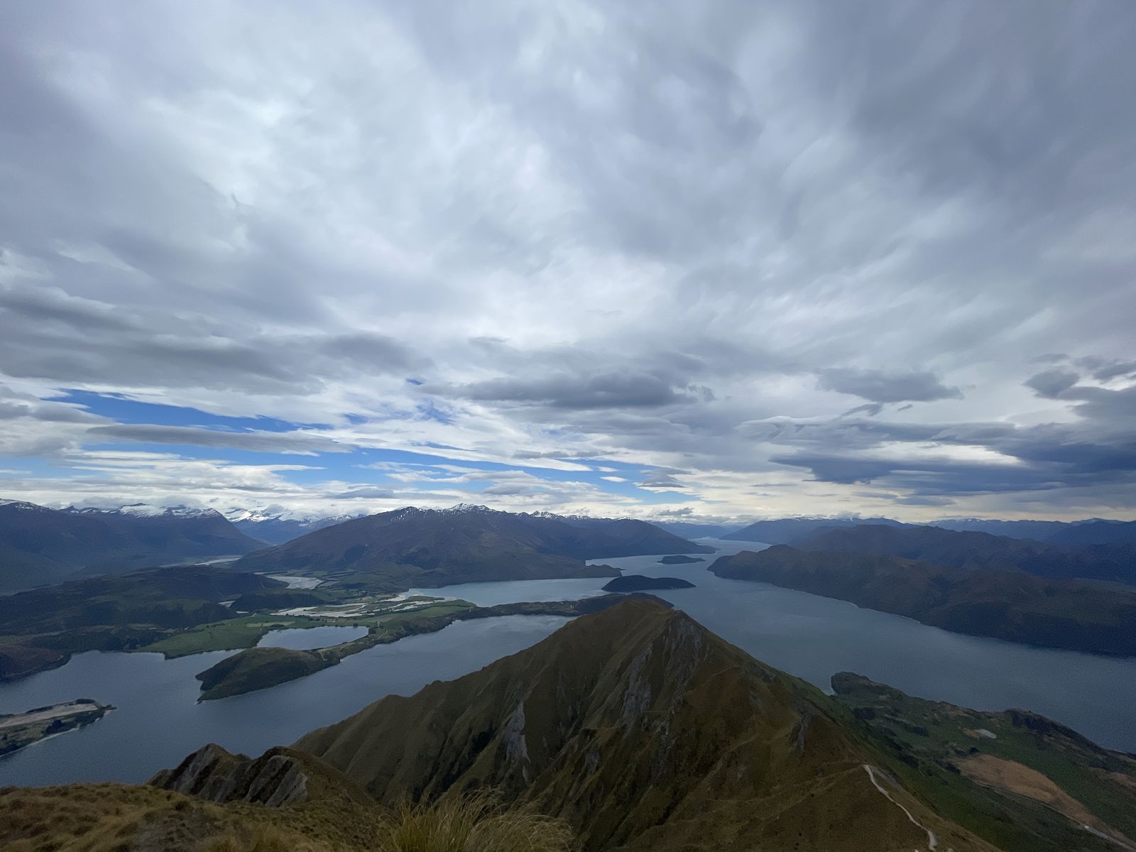 The ridge of Roys Peak above Lake Wānaka and its islands, with mountains under a cloudy sky