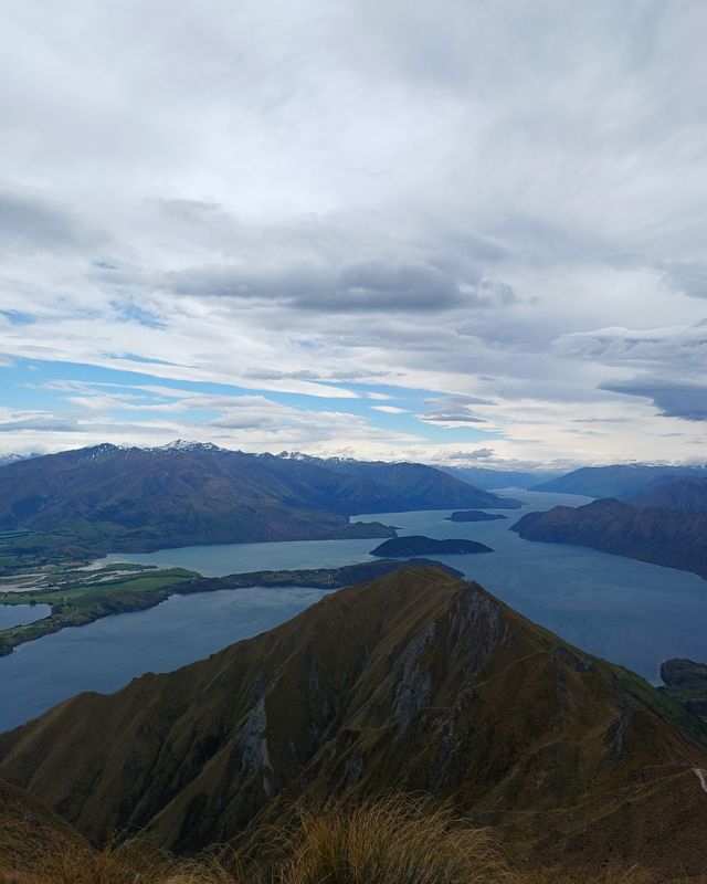 The ridge of Roys Peak above Lake Wānaka and its islands, with mountains under a cloudy sky